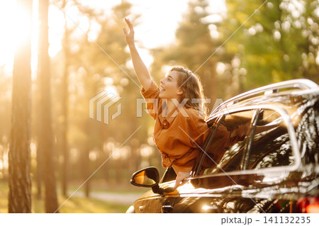 A young woman feels freedom leaning out of a car window in a sunny forest. Travel concept. A young woman feels freedom leaning out of a car window in a sunny forest. Travel concept. 141132235
