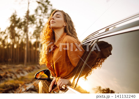 A young woman feels freedom leaning out of a car window in a sunny forest. Travel concept. A young woman feels freedom leaning out of a car window in a sunny forest. Travel concept. 141132240