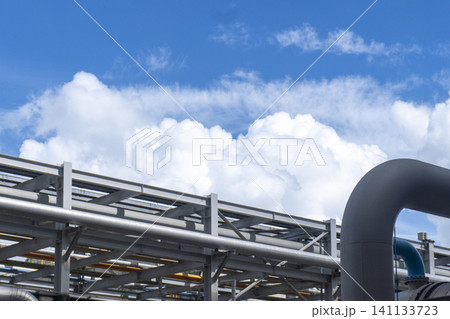 Industrial metal pipelines and steel framework at a petrochemical or gas plant against a bright blue sky with white clouds representing modern industrial infrastructure. Industrial metal pipelines and steel framework at a petrochemical or gas plant against a bright blue sky with white clouds representing modern industrial infrastructure. 141133723