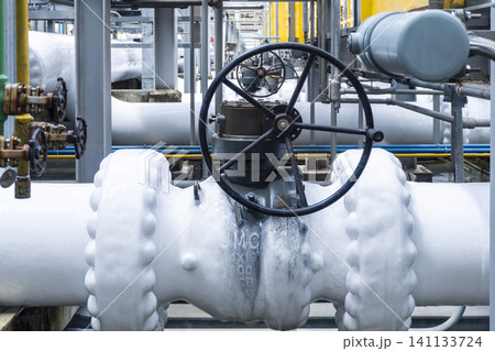 Close-up of a large manual gate valve on frozen pipelines covered in ice due to cryogenic gas flow inside an LNG or industrial plant surrounded by yellow-painted steel structures. 141133724