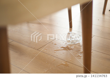 A broken glass spills liquid onto a wooden floor, creating a mess 141134763