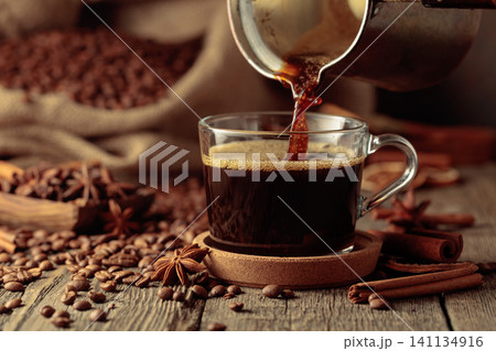 Black coffee is poured from a copper coffee pot into a glass cup. 141134916