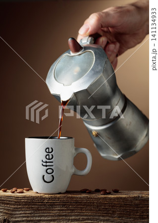 Black coffee is poured from a classic Italian aluminium moka pot into a coffee cup. Black coffee is poured from a classic Italian aluminium moka pot into a coffee cup. 141134933