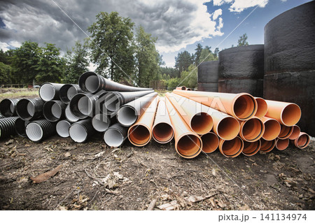 Several pipes are arranged in piles beside large tanks in a construction area. The cloudy sky hints at possible rain later in the day Several pipes are arranged in piles beside large tanks in a construction area. The cloudy sky hints at possible rain later in the day 141134974