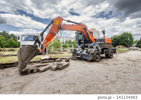 A large excavator with an orange boom lifts large rocks during construction in the city. The sun is shining, but the sky is cloudy A large excavator with an orange boom lifts large rocks during construction in the city. The sun is shining, but the sky is cloudy 141134983