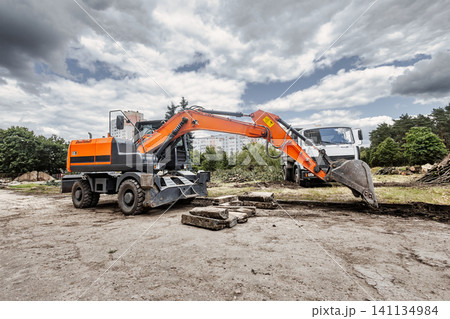 An excavator is working at a construction site, moving soil and rocks, and a truck is parked nearby in a busy area with many trees 141134984