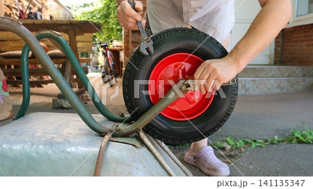 A man repairs an overturned wheelbarrow. Using two wrenches, he tightens the axle lugs of a new black wheel with a bright red rim. The work takes place outside near the house. A man repairs an overturned wheelbarrow. Using two wrenches, he tightens the axle lugs of a new black wheel with a bright red rim. The work takes place outside near the house. 141135347