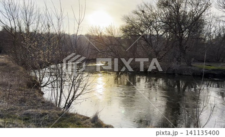 View on calm river in early spring reflects soft sunlight. Warm rays shimmer on water surface, creating peaceful at sunset. The image conveys tranquility and seasonal awakening in nature. Slow motion View on calm river in early spring reflects soft sunlight. Warm rays shimmer on water surface, creating peaceful at sunset. The image conveys tranquility and seasonal awakening in nature. Slow motion 141135400