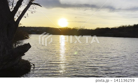 Serene lake bathed in warm sunset light with golden reflections shimmering across the calm water. The surrounding landscape softly illuminated, creating peaceful and tranquil summer evening atmosphere Serene lake bathed in warm sunset light with golden reflections shimmering across the calm water. The surrounding landscape softly illuminated, creating peaceful and tranquil summer evening atmosphere 141136259
