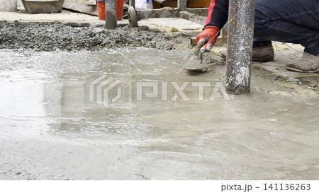 Male hands in protective gloves uses trowel to spread and level fresh cement in yard. Material is carefully applied to surface as part of preparation work before it hardens into concrete layer Male hands in protective gloves uses trowel to spread and level fresh cement in yard. Material is carefully applied to surface as part of preparation work before it hardens into concrete layer 141136263