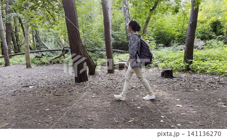 Young girl going along path at summer forest. Female hiker with backpack walking among trail at wild nature. The scene reflecting adventure, travel and connection with wild nature. Slow motion Young girl going along path at summer forest. Female hiker with backpack walking among trail at wild nature. The scene reflecting adventure, travel and connection with wild nature. Slow motion 141136270