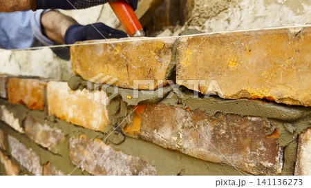 Adult male hands in protective gloves apply cement with a trowel and place bricks to build brick wall for house. The worker carefully spreads mortar and sets each brick during the construction process Adult male hands in protective gloves apply cement with a trowel and place bricks to build brick wall for house. The worker carefully spreads mortar and sets each brick during the construction process 141136273