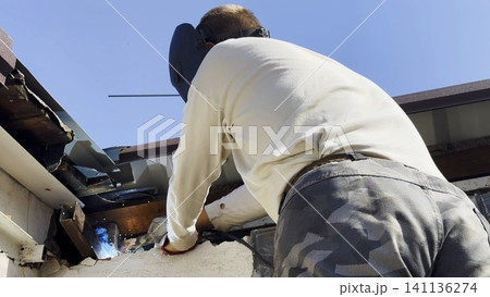 Manual laborer in protective mask welds iron detail at construction. Professional young repairman with welding machine working at the site. Male welder operating with metal on house. Close up Manual laborer in protective mask welds iron detail at construction. Professional young repairman with welding machine working at the site. Male welder operating with metal on house. Close up 141136274