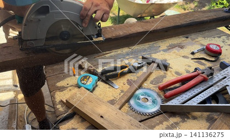 Arms of professional repairman sawing wooden construction with a circular saw at construction. Male hands of adult craftsman cuts wood using electric grind wheel at the site. Slow motion Arms of professional repairman sawing wooden construction with a circular saw at construction. Male hands of adult craftsman cuts wood using electric grind wheel at the site. Slow motion 141136275