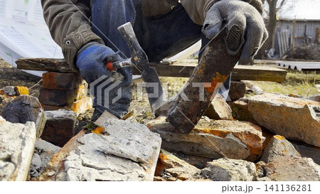 Male hands in protective gloves uses hammer to remove old cement from used bricks during renovation work. The process involves carefully cleaning brick surface to prepare it for reuse in construction. Male hands in protective gloves uses hammer to remove old cement from used bricks during renovation work. The process involves carefully cleaning brick surface to prepare it for reuse in construction. 141136281