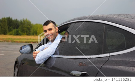 Handsome successful entrepreneur in shirt leaning out of driver window of modern electric car and looking into camera. Portrait of smiling businessman in innovative electrical vehicle at rainy day Handsome successful entrepreneur in shirt leaning out of driver window of modern electric car and looking into camera. Portrait of smiling businessman in innovative electrical vehicle at rainy day 141136283