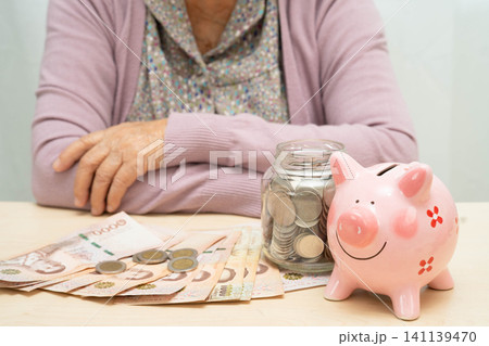 Retired elderly woman counting coins money with piggy bank and worry about monthly expenses and treatment fee payment. Retired elderly woman counting coins money with piggy bank and worry about monthly expenses and treatment fee payment. 141139470