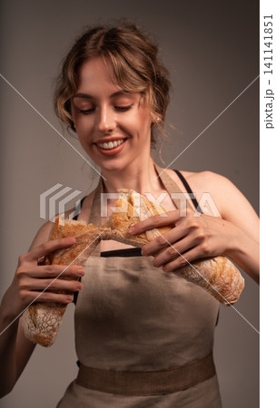 Smiling woman wearing an apron breaking fresh baguette, celebrating baking and culinary enjoyment Smiling woman wearing an apron breaking fresh baguette, celebrating baking and culinary enjoyment 141141851