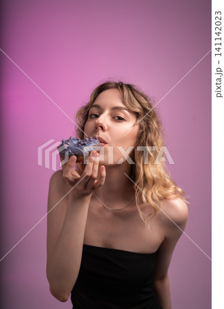 Young woman enjoying sweet donut with purple frosting and sprinkles on a vibrant pink background Young woman enjoying sweet donut with purple frosting and sprinkles on a vibrant pink background 141142023