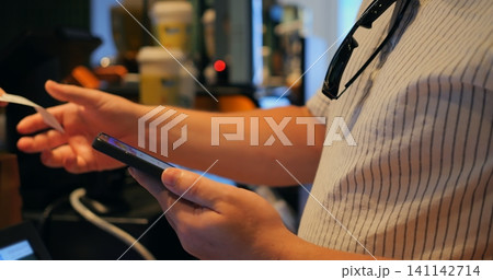 Person holding smartphone and payment card for mobile payment at checkout counter, close view of hands, phone screen, and pinstripe shirt, blurred cafe background visible behind payment scene 141142714