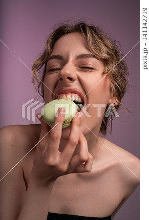 Young woman enjoying a sweet treat, taking a large bite of a green macaron with eyes closed 141142719