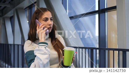 Indoor bridge walk with lively atmosphere. Casual woman multitasking on urban bridge during daylight. City woman engages in call while strolling indoors with coffee and natural light Indoor bridge walk with lively atmosphere. Casual woman multitasking on urban bridge during daylight. City woman engages in call while strolling indoors with coffee and natural light 141142730