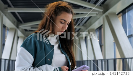 Closeup young woman texting on smartphone, wearing green and white jacket, soft window light illuminating face, absorbed expression while scrolling messages, handheld device and attentive posture Closeup young woman texting on smartphone, wearing green and white jacket, soft window light illuminating face, absorbed expression while scrolling messages, handheld device and attentive posture 141142733