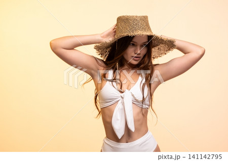 Woman wearing white high-waisted bikini and straw hat, posing against a warm background for summer fashion 141142795