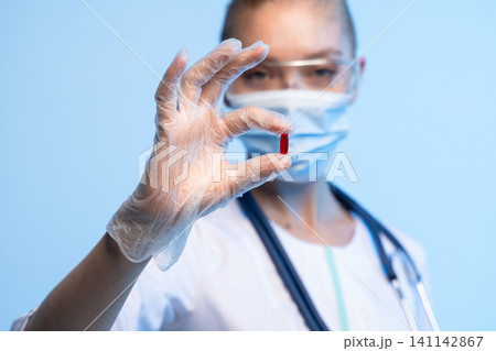 Healthcare worker in mask, gloves, and glasses holding a single red capsule, symbolizing medicine, research, and therapy Healthcare worker in mask, gloves, and glasses holding a single red capsule, symbolizing medicine, research, and therapy 141142867