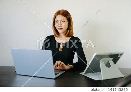 Young professional Asian woman multitasking on laptop and tablet in a modern workspace, showcasing productivity, digital work, and business lifestyle. Young professional Asian woman multitasking on laptop and tablet in a modern workspace, showcasing productivity, digital work, and business lifestyle. 141143226