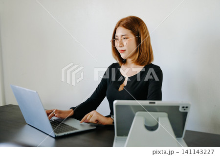 Young professional Asian woman multitasking on laptop and tablet in a modern workspace, showcasing productivity, digital work, and business lifestyle. Young professional Asian woman multitasking on laptop and tablet in a modern workspace, showcasing productivity, digital work, and business lifestyle. 141143227