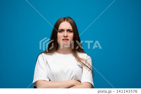 Young woman reacting with disgust or displeasure, frowning with arms crossed against a blue studio background Young woman reacting with disgust or displeasure, frowning with arms crossed against a blue studio background 141143576
