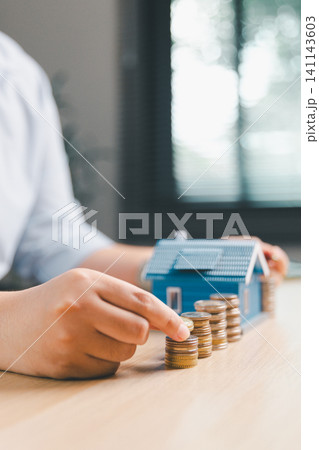 Close-up hand adding coin to stacked coins with blue house model in background, representing home savings growth, mortgage deposit, and real estate investment plan. 141143603