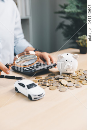 Business person using magnifying glass to review car savings, piggy bank, calculator, coins, and white toy car, representing auto loan analysis and vehicle finance planning. 141143608