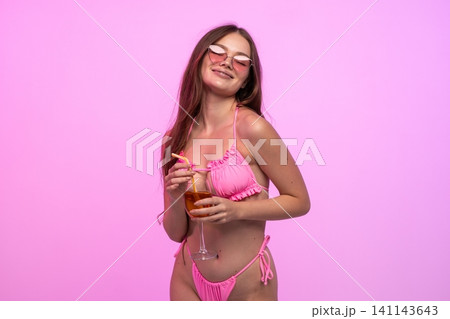 Young woman enjoying summer vacation, wearing a pink bikini and heart-shaped sunglasses, holding a refreshing cocktail Young woman enjoying summer vacation, wearing a pink bikini and heart-shaped sunglasses, holding a refreshing cocktail 141143643
