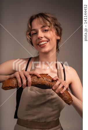 Smiling woman wearing an apron breaking a fresh whole grain baguette, enjoying warm homemade bread Smiling woman wearing an apron breaking a fresh whole grain baguette, enjoying warm homemade bread 141144452