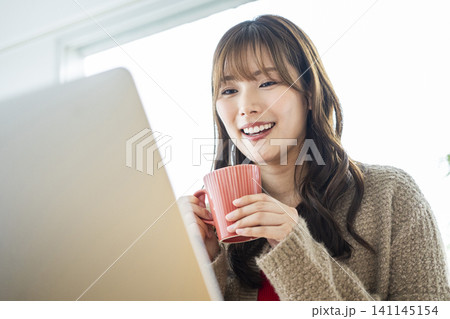 コーヒーを飲みながらパソコンで仕事をする若い女性 コーヒーを飲みながらパソコンで仕事をする若い女性 141145154