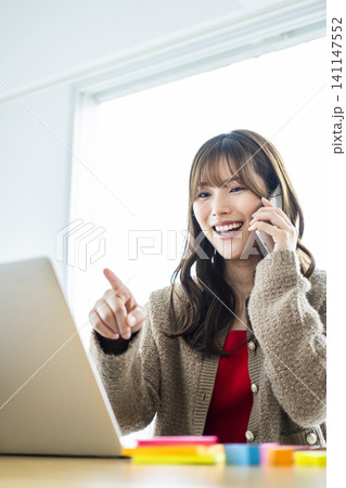 パソコンを見ながらスマホで打ち合わせをする若い女性 パソコンを見ながらスマホで打ち合わせをする若い女性 141147552