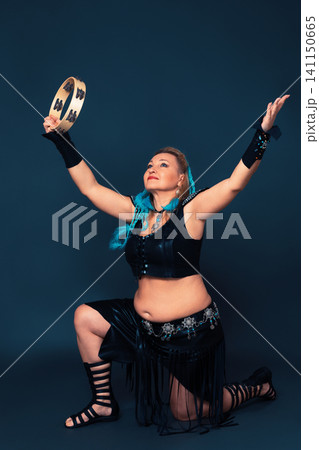 Female dancer stands on one knee with her arms raised up, dances with tambourine and looks up. Mature woman dressed in handmade performance costume - leather top, skirt, bracers, gladiator boots 141150665