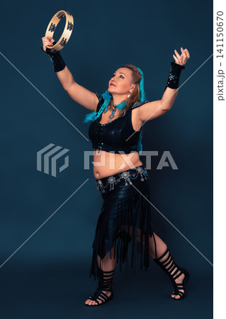 Confidence woman dancing with tambourine, contributing to dynamic ambiance. Curvy female dancer is dressed in black, handmade costume, which includes leather top and skirt, bracers, gladiator boots Confidence woman dancing with tambourine, contributing to dynamic ambiance. Curvy female dancer is dressed in black, handmade costume, which includes leather top and skirt, bracers, gladiator boots 141150670