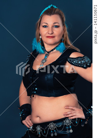 Woman belly dancer wearing leather costume with blue feathers and silver jewelry. Portrait of blonde bellydancer with arms akimbo and looking at camera Woman belly dancer wearing leather costume with blue feathers and silver jewelry. Portrait of blonde bellydancer with arms akimbo and looking at camera 141150671