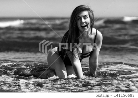 Woman laughing in ocean water at beach. Happy lady in swimsuit kneeling in sea waves. Cinematic black and white portrait with film grain and retro aesthetic. Summer vacation and lifestyle concept Woman laughing in ocean water at beach. Happy lady in swimsuit kneeling in sea waves. Cinematic black and white portrait with film grain and retro aesthetic. Summer vacation and lifestyle concept 141150685