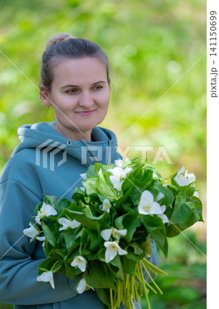 Female person in teal hoodie holding fresh trillium blooms with lush green leaves. Soft natural lighting, shallow depth of field, serene outdoor mood. Ideal for nature, wellness, or botanical themes Female person in teal hoodie holding fresh trillium blooms with lush green leaves. Soft natural lighting, shallow depth of field, serene outdoor mood. Ideal for nature, wellness, or botanical themes 141150699