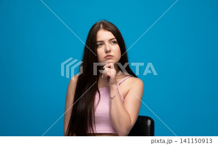 Woman in pink top looking up with a thoughtful expression, contemplating and questioning against a vibrant blue studio backdrop 141150913