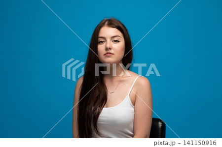 Young woman with long brunette hair and confident expression, sitting against a vibrant blue studio background Young woman with long brunette hair and confident expression, sitting against a vibrant blue studio background 141150916