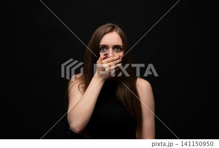 Young woman covering her mouth with her hand, expressing shock and fear with wide eyes on a dark background Young woman covering her mouth with her hand, expressing shock and fear with wide eyes on a dark background 141150950
