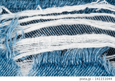Close up view of blue denim fabric with distressed ripped texture. Worn jeans material with frayed white cotton threads. Textile pattern for fashion design and clothing textured background Close up view of blue denim fabric with distressed ripped texture. Worn jeans material with frayed white cotton threads. Textile pattern for fashion design and clothing textured background 141151044
