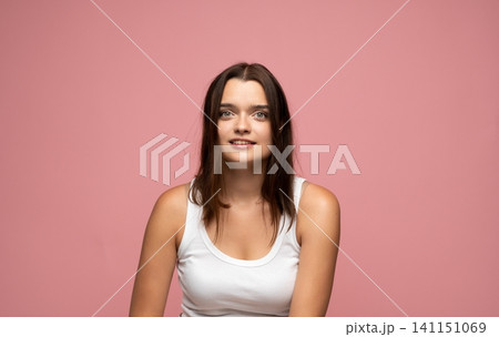 Young brunette woman looking at camera with friendly expression, wearing a white tank top on a pink background Young brunette woman looking at camera with friendly expression, wearing a white tank top on a pink background 141151069