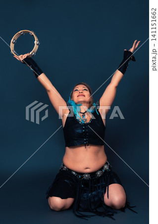 Happy woman dancer is kneeling and raising her arms up, holding tambourine in one of them Happy woman dancer is kneeling and raising her arms up, holding tambourine in one of them 141151662