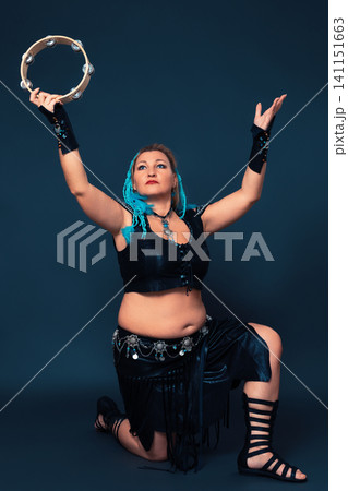Dancer assumes position with one knee raised, arms extended upward, performs dance with tambourine Dancer assumes position with one knee raised, arms extended upward, performs dance with tambourine 141151663
